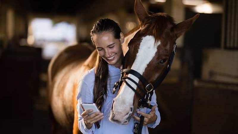 Junge Frau schaut lächelnd auf ihr Smartphone. Sein Pferd schaut ihm über die Schulter.