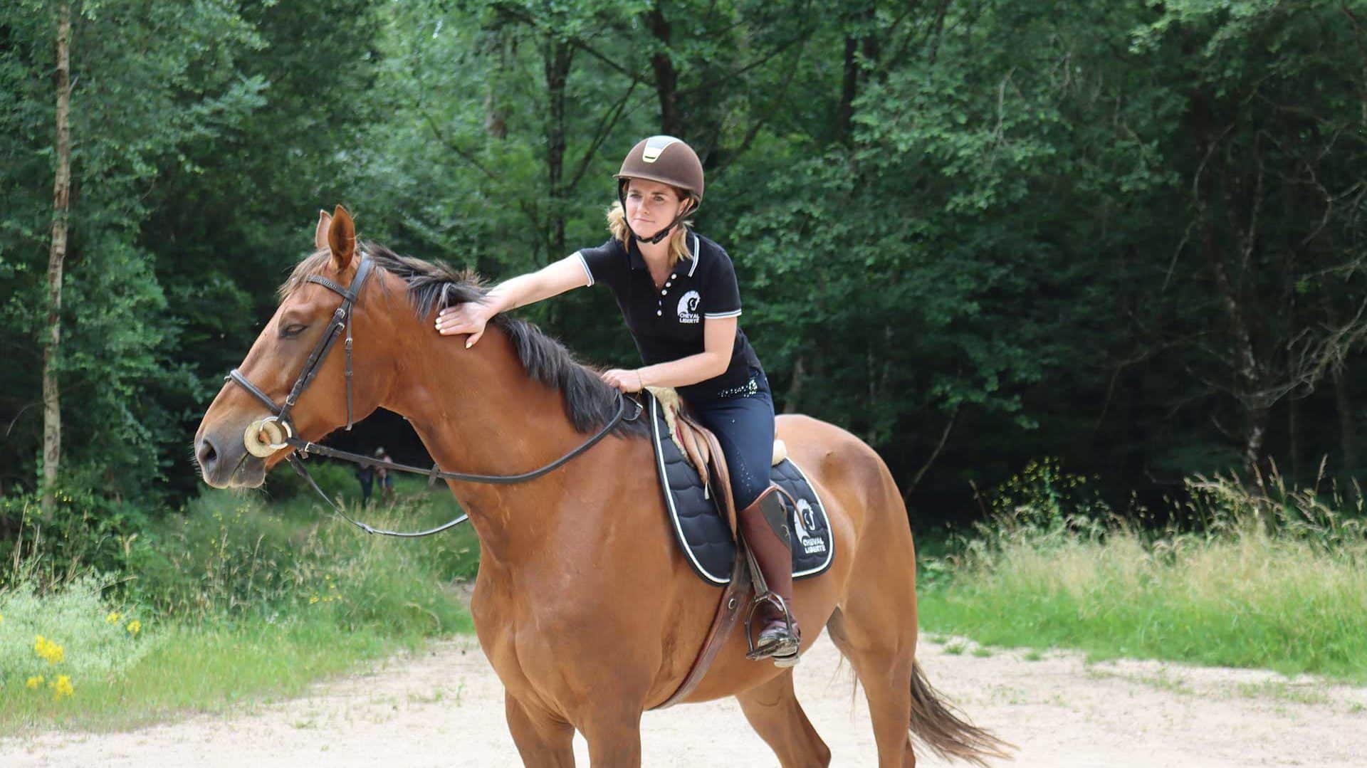 Eine stehende Reiterin streichelt ihr Pferd im Wald und lächelt.
